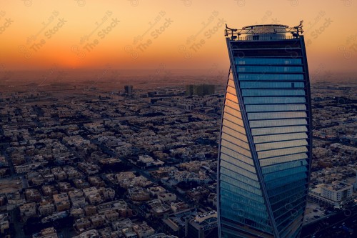 A shot showing the scheduled tower, one of the buildings and landmarks of the city of Riyadh, in front of it, a group of residential houses, and the sky is clear in orange at sunset.