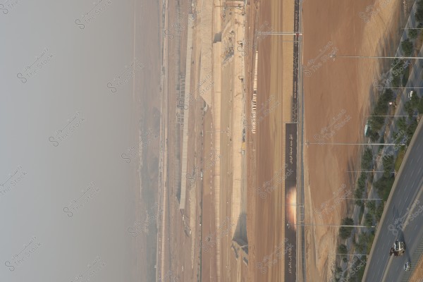 صورة لشارع واسع مع طريق مسفلت إلى اليمين وبعض السيارات والدراجات النارية تسير عليه. يظهر على الجانب الأيسر منطقة قيد الإنشاء مع تكديس كبير من التربة والحجارة وآليات ثقيلة تعمل في الموقع. هناك أيضًا لافتة كبيرة مكتوب عليها باللغة العربية, ومثبتة على حاجز معدني يفصل بين منطقة البناء والطريق.