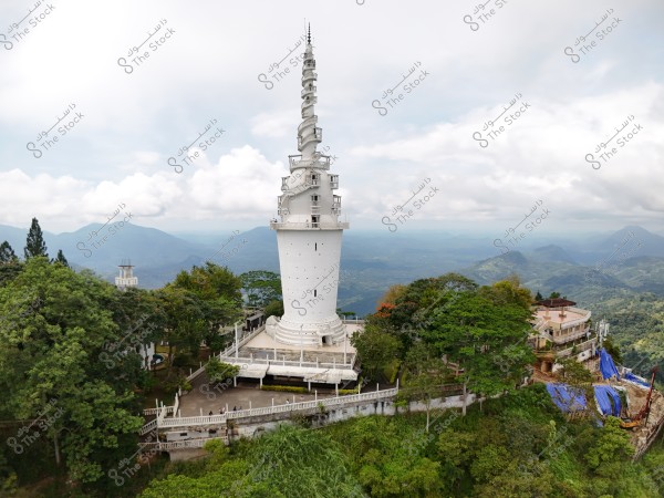 صورة تظهر برجًا أبيض طويلًا بقاعدة دائرية, يبدو كأنه منارة أو برج مراقبة, مُحاط بالغابات الخضراء والمناظر الطبيعية الجبلية في الخلفية. يمكن رؤية السماء ملبدة بالغيوم, مما يضفي جمالية كبيرة على المشهد.