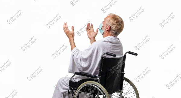 A man sitting in a wheelchair wearing a face mask. He appears to be wearing a white robe and has his hands raised in a gesture of prayer or meditation. The background is white.