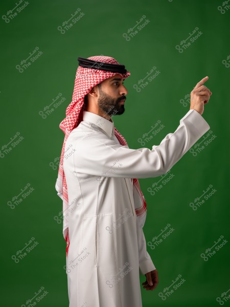 رجل يرتدي الثوب السعودي التقليدي مع الشماغ يقف على خلفية خضراء ويرفع يده.