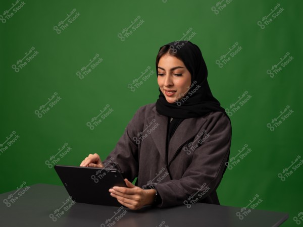 امرأة ترتدي حجابًا وتجلب معطفًا وهي تستخدام جهاز لوحي أمام خلفية خضراء.