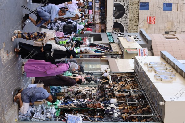 An image of a busy street market with people shopping near a store displaying a variety of shoes neatly arranged on shelves. The shoppers are wearing diverse clothing, including abayas, hijabs, and colorful jackets. Storefronts with Arabic signs are visible in the background, indicating a local shopping area.