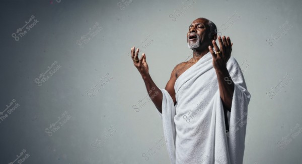 An image of an elderly person wearing a white garment resembling the attire used in Hajj and Umrah. The person stands with hands raised upwards, with an expression of contemplation or prayer, against a gray background.