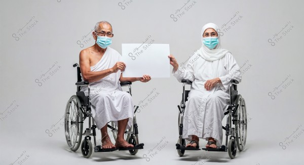 The image shows an elderly man and woman seated in wheelchairs. The man is wearing traditional white ihram clothing, and the woman is dressed in a white abaya and hijab. Both are wearing blue face masks and are holding a blank white sign between them. The background is plain white.