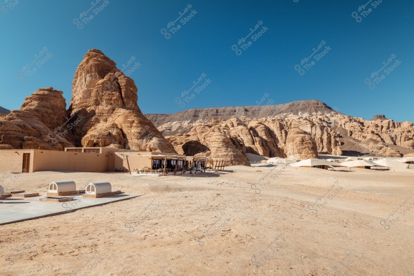 A desert landscape featuring striking rock formations under a clear blue sky in the AlUla region of Saudi Arabia. There is a modern architectural structure that blends with the surroundings, featuring canopies and shadows, with sandstone used in construction. Sand dunes and large rocks surround the buildings.