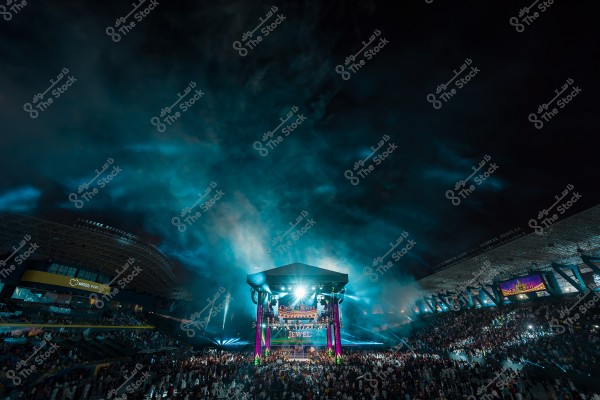 مشهد لملعب مزدحم خلال حدث كبير في الليل, مع أضواء ساطعة ودخان يملأ الجو حول المسرح الرئيسي.