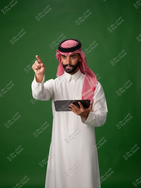 An image of a man wearing traditional Saudi white thobe and red shemagh with a black agal, standing in front of a green background. He is holding a tablet in his hands and appears to be pointing forward with his other hand in a serious manner.