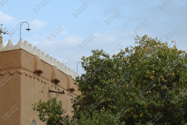 The image shows a section of a traditional brown building adorned with white crenellations on top, alongside dense green trees. The sky is blue with some clouds.
