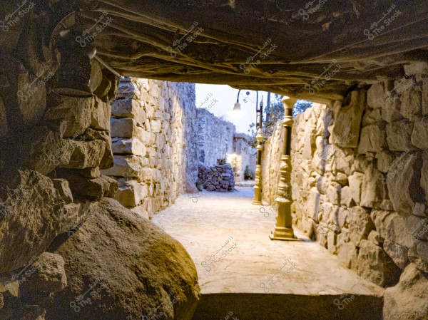 A stone walkway covered with wooden ceilings, leading to another passageway lined with elegant stone walls and lit by classic street lamps. In the background, additional stone walls are visible, along with an open area with plants.