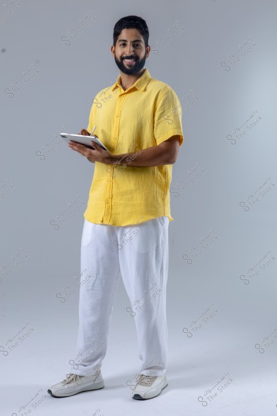 A portrait of a man standing and smiling while holding a tablet. He is wearing a yellow shirt and white pants, paired with white sneakers. The background is a neutral gray. The man appears in casual, comfortable attire that might be inspired by modern Arab fashion.