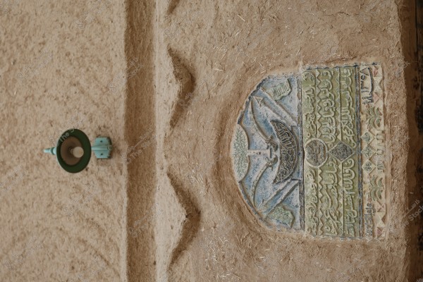 نقش عربي محفور على جدار طيني. يظهر التصميم الفني التقليدي عناصر زخرفية متعددة مثل السيوف وأشجار النخيل, وتحيط بها نقوش عربية. على الجانب الأيسر من الصورة, يوجد مصباح قديم باللون الأخضر الفاتح مثبت على الحائط, مما يضفي طابعاً تقليدياً على المشهد.