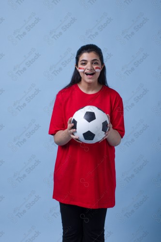 Portrait of a Saudi woman wearing a red T-shirt cheering the football team on a blue background and showing expressions of joy and enthusiasm, World Cup.