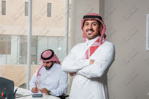 رجل سعودي يرتدي الزي السعودي التقليدي يقف مكتوف اليدين أثناء إجتماع في العمل نهارًا