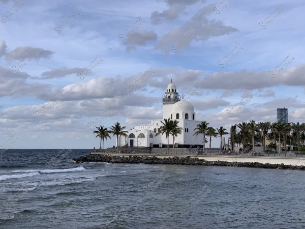 Image of a large mosque with a white dome and a minaret overlooking a waterfront lined with palm trees along a promenade. There are sea waves in the foreground and a blue sky with scattered clouds in the background. A tall building is also visible on the right side in the background.