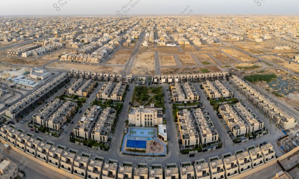 An aerial view of a modern residential neighborhood in a desert area, featuring a collection of villas and neatly arranged houses, with narrow streets separated by green spaces and sports courts. These houses are spread in a regular pattern surrounded by undeveloped open areas, showcasing a blend of urban development and the surrounding desert landscape.