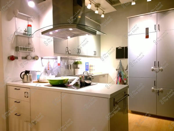 Image of a modern, sleek kitchen with an open design. The kitchen features a central white island with a built-in stovetop and a large green bowl. There are wall-mounted shelves holding various kitchen utensils, including a silver kettle and a glass container. The kitchen is illuminated by multiple ceiling lights and has white cabinets with a light wood floor. There are also pots and pans placed on the countertop.