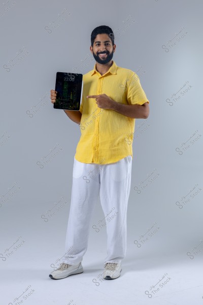 A man stands smiling in a studio with a grey background, holding a tablet in one hand and pointing at it with the other. He is wearing a yellow shirt, white trousers, and white sneakers. The image conveys a modern and casual impression.