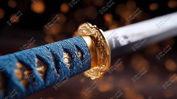 Close-up image of a samurai sword hilt, showing intricate details on the handle wrapped in blue cord with golden accents. The handle is connected to a highly decorated golden guard. The background is blurred with mysterious orange circular bokeh lights.