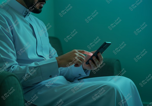 The image shows a man sitting on a sofa wearing traditional white attire, likely from the Gulf region such as Saudi Arabia. He is using a smartphone with one hand in dim lighting.