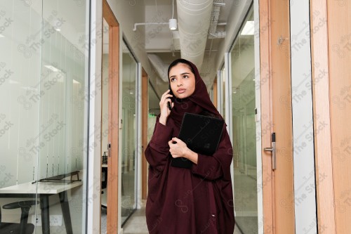 Saudi woman wearing an abaya stands in the meeting room in front of the glass front, holding a tablet ، during the day