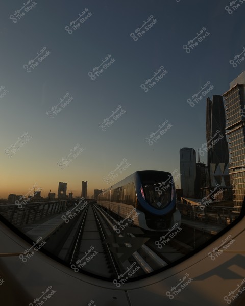 An image of a metro train traveling on elevated tracks in an urban cityscape at sunset. The train has a modern design with a large glass front. In the background, various skyscrapers are visible, reflecting the golden rays of the sun. The sky is clear with warm tones indicating the end of the day.