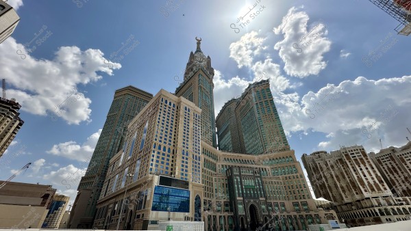 An image of the Abraj Al Bait complex in Mecca, Saudi Arabia, prominently featuring the main tower known as the Makkah Clock Tower in the center. The towers are painted in a combination of gold and blue colors, showcasing their modern and traditional architectural features. The sky is blue with soft white clouds, and the sun shines brightly above.
