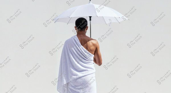 An image of a person from behind holding a white umbrella. The person appears to be wearing a white wrap similar to Ihram clothing, standing against a white background. The scene is simple and calm.