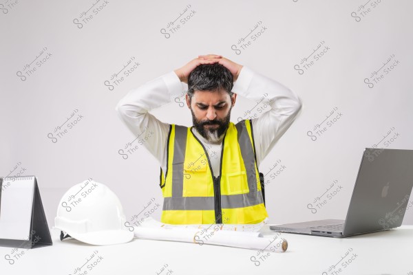 The image shows a man sitting behind a desk wearing a yellow reflective vest, suggesting he might be an engineer or construction worker. In front of him are a white helmet, architectural blueprint papers, and a laptop. The man has his hands on his head and appears to be concerned or stressed. The background is white and clean.