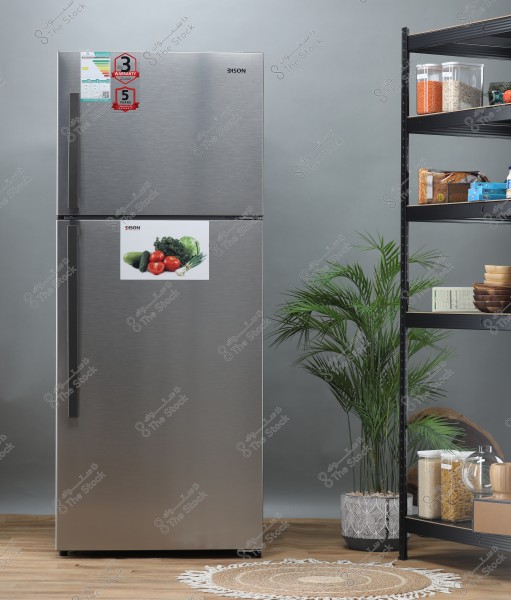 A photo of a silver metallic refrigerator with a vegetable sticker on its door featuring tomatoes, cucumber, and other vegetables. The refrigerator has red stickers indicating warranty periods of 3 and 5 years. To the right, there are shelves holding various food containers such as lentils, grains, and bread. On the floor, there is a small round rug and green plants decorating the area.