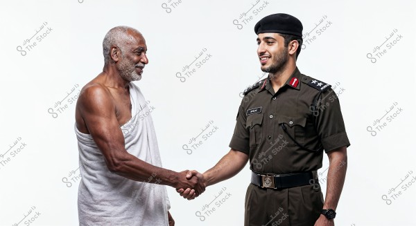 The image shows two men shaking hands. The man on the left appears to be elderly and is wearing traditional white Ihram clothing. The man on the right is wearing a military uniform with a black cap, possibly from Saudi Arabia, and looks young. The background is plain white.