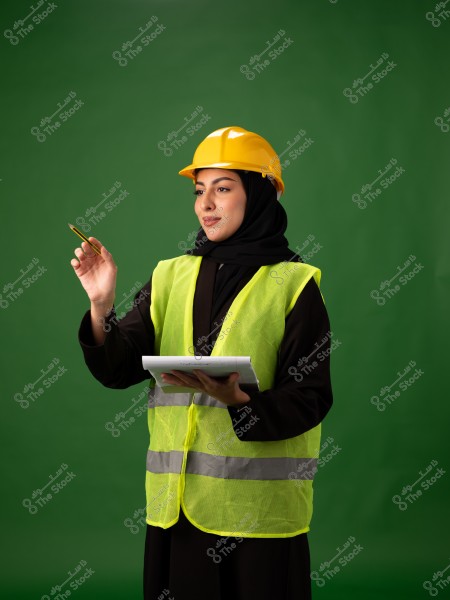 A woman wearing a black hijab, a yellow hard hat, and a yellow reflective vest. She is holding a notebook and a pen, standing in front of a green background. She appears to be working in construction or engineering.