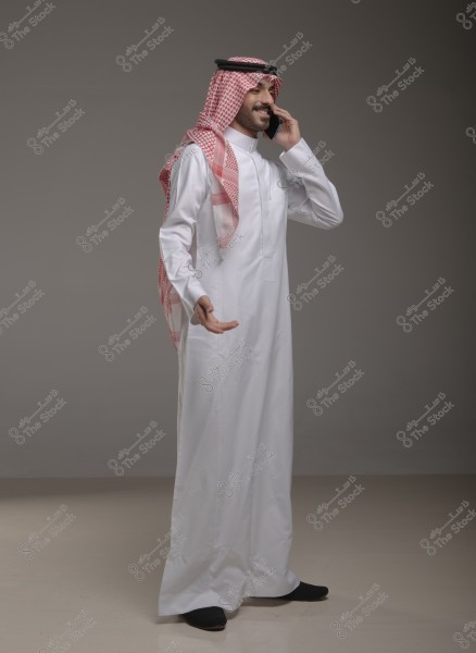 A photo of a man wearing traditional Saudi attire, consisting of a white thobe, a red checkered headscarf (ghutra), and a black agal. He is standing, smiling, and talking on a mobile phone. The background is plain and gray.