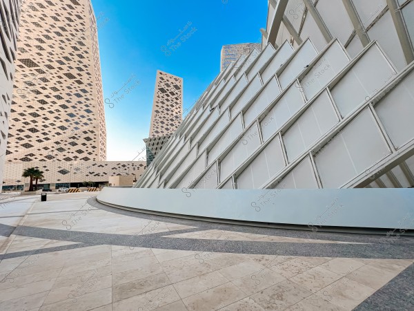 صورة لمبانٍ حديثة ذات تصميم معماري مميز في بيئة حضرية. تظهر المباني بواجهات مغطاة بنمط شبكي أبيض يحتل جانبًا كبيرًا من الصورة تحت سماء زرقاء صافية. توجد أشجار نخيل في الجزء الأيسر من الصورة, مما يعطي إشارات لمنطقة ذات مناخ صحراوي.