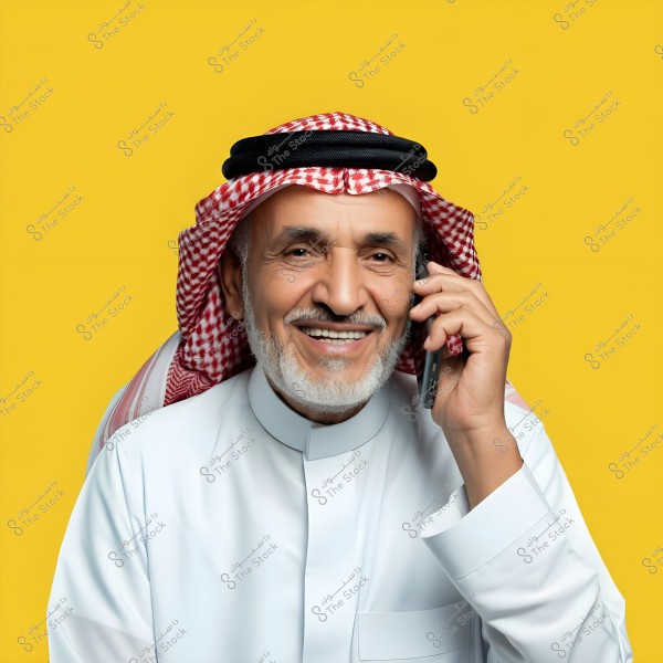 Portrait of an elderly man wearing a white traditional Arabic thobe and a red and white ghutra on his head, smiling while talking on the phone. The background is bright yellow.
