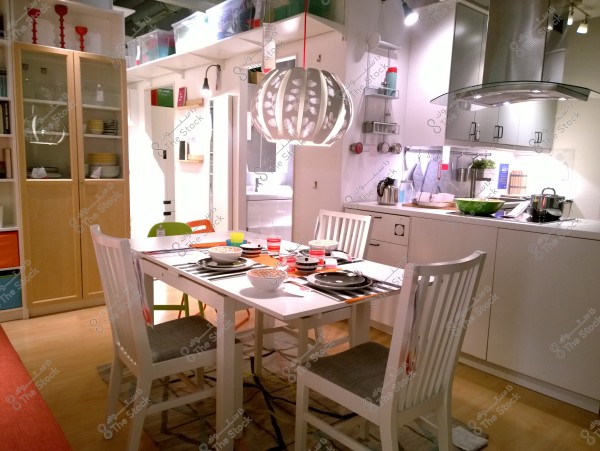 The image shows a modern and elegantly lit kitchen with a white dining table surrounded by white chairs. The table is set with colorful dishes and bowls. A decorative hanging lamp is above the table. In the background, there are white cabinets with neatly stacked dishes and kitchen utensils like a kettle and a pot.