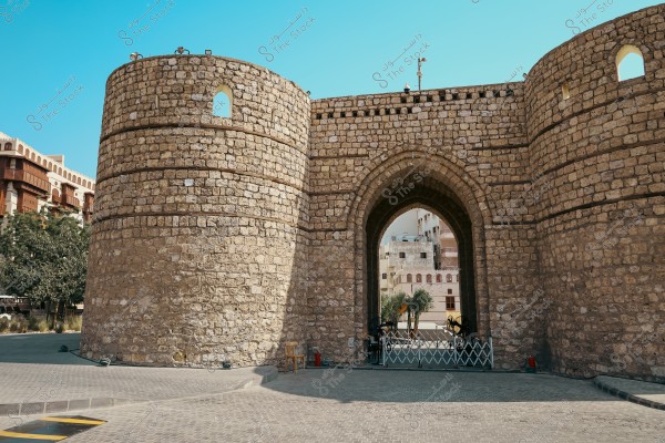 An image showing a historic stone gate in the city of Jeddah, Saudi Arabia. The gate features a large arch in the center and thick stone walls. In the background, modern apartment buildings and palm trees are visible. The sky is clear and blue, adding a touch of heritage and antiquity to the scene.