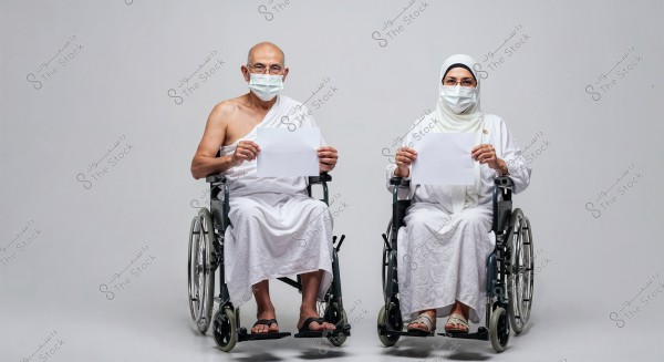 A man and a woman seated in wheelchairs, wearing white Ihram clothing and medical masks on their faces. Each is holding a blank white sheet of paper. The background is plain white.