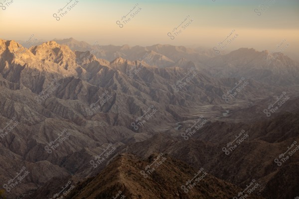 Panoramic image of sprawling mountains with rugged terrain under soft sunlight. The mountains are depicted in dark brown and gray hues, with gradient layers receding into the misty horizon. A narrow valley runs between the mountains, with a faint glimmer in the center of the scene.
