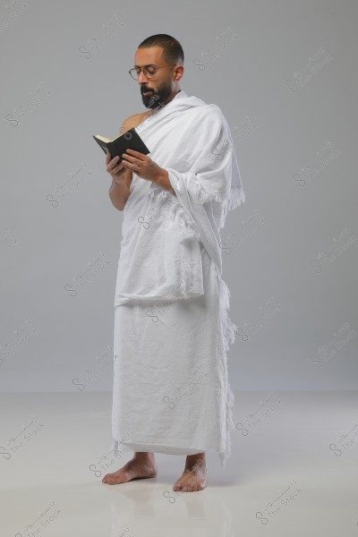 Image of a man standing against a gray background, wearing a white Ihram garment and holding a small book as he reads, appearing to be in contemplation or worship. The man is barefoot and wearing glasses.