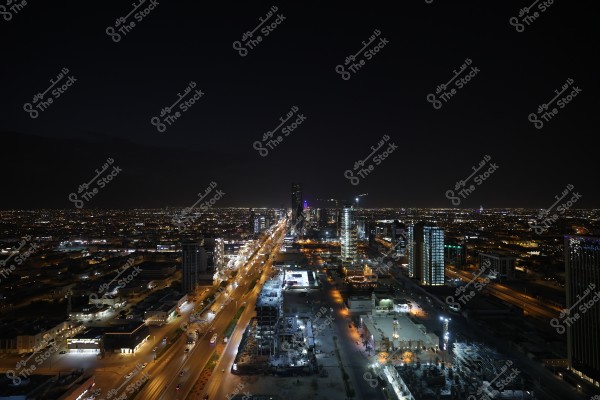 Nighttime aerial view of a modern city with a brightly lit road stretching through the center. Tall buildings illuminate the skyline amidst a vast urban area, and city lights extend to the horizon in the distance.