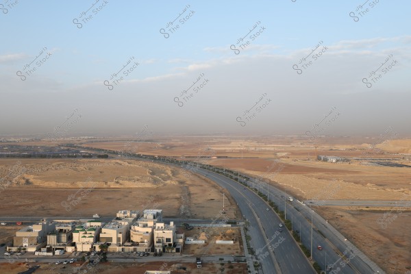 Aerial view of a city with several modern architectural buildings in the foreground surrounded by a vast, arid desert area. A long road runs parallel to the buildings, with a few cars visible on the road. The sky is blue with some light clouds at the edges.