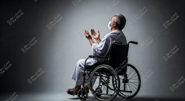 An elderly man sits in a wheelchair wearing an Ihram in a shaded setting. He raises his hands in a praying position. He is wearing a medical face mask.