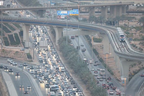 The image shows an urban scene of a modern city with a busy highway filled with cars under an elevated bridge. On the bridge, a modern train is seen running on its track. Greenery is distributed along the sides of the road, and there are directional signs with information in both Arabic and English. The image highlights features of advanced infrastructure.