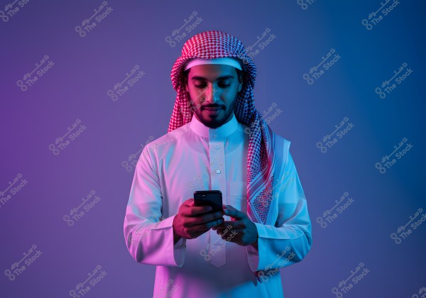 A portrait of a man wearing a white traditional thobe and a red and white checked headscarf, suggesting Saudi Arabian attire. He is standing and using a mobile phone, with blue and pink colored lighting creating a modern and vibrant effect.