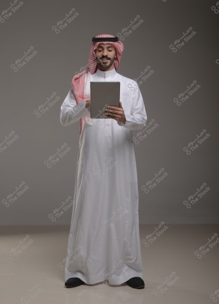 صورة لرجل يرتدي ثوباً أبيض وغترة حمراء مع عقال, يمسك جهازًا لوحيًا بيده الاثنين ويبتسم. يبدو أن الصورة تمثل بورتريه لشخصية من المملكة العربية السعودية.