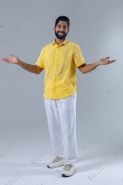 A portrait of a man standing and smiling, wearing a short-sleeved yellow shirt and white trousers with light-colored sneakers. The man is standing against a gray background, appearing to be in a studio. His hands are open to his sides, giving an impression of welcoming or presenting.