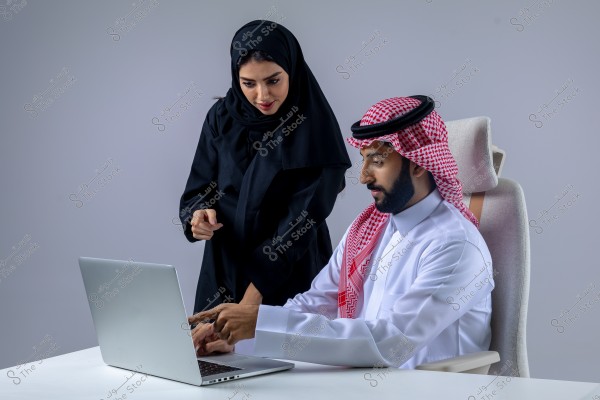 An image of a man and woman working together in an office setting. The man is seated on a chair using a laptop, wearing a white thobe and a red and white ghutra with a black agal. The woman stands beside him, pointing at the computer screen, dressed in a black abaya and hijab. The background is simple and grey.