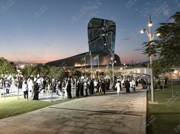 A large gathering of people in an open square in front of a distinctive modern building, likely the King Abdulaziz Center for World Culture in Dhahran, Saudi Arabia, also known as \"Ithra.\" The building features a sleek, modern design, while several Saudi flags are flying in the foreground. Most attendees are dressed in traditional Arabic clothing, such as white thobes for men and black abayas for women. The scene suggests a cultural or social event taking place in the evening with a beautiful sunset sky in the background.