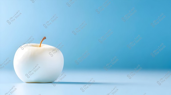 An image of a white apple placed on a blue surface with a light blue background. The apple has a short brown stem and a glossy appearance.